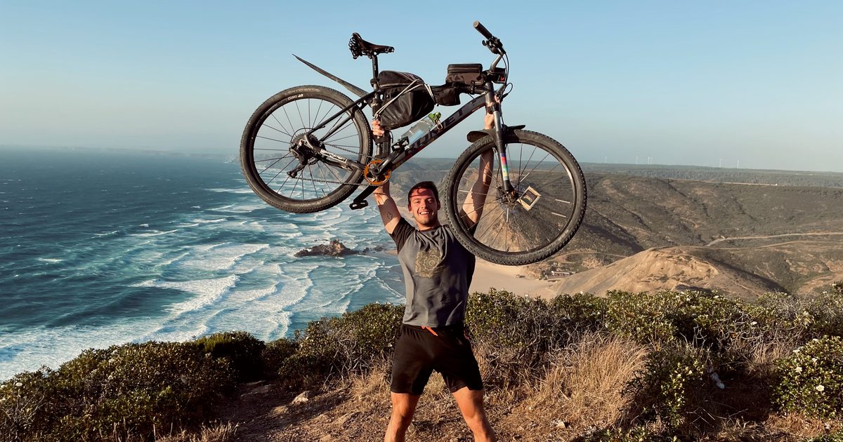 Cyclist lifting a loaded gravel bike overhead on a clifftop above the Atlantic, Algarve west coast