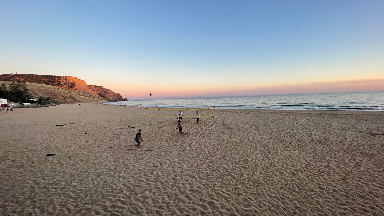 People playing beach volleyball on a wide sandy beach at dusk, with cliffs in the distance